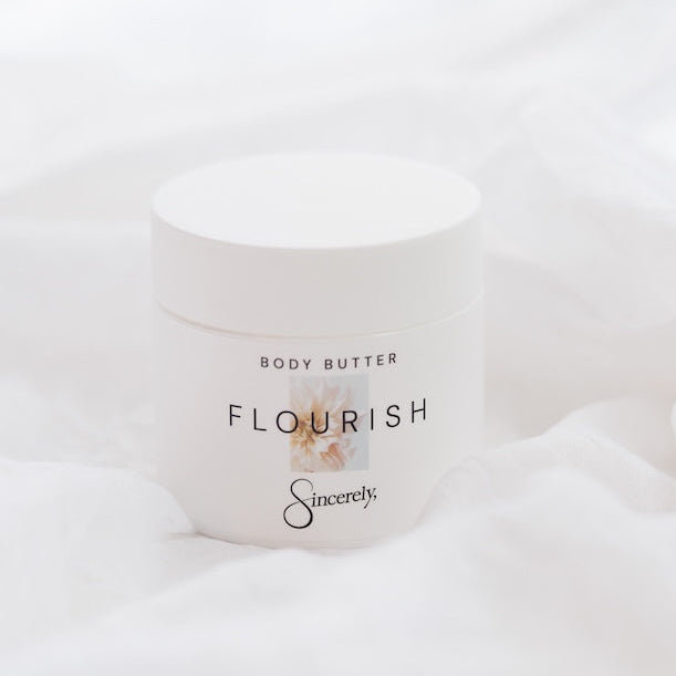A white jar of Sincerely body butter labeled 'Flourish' resting on soft white draped fabric.