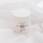 A white jar of Sincerely body butter labeled 'Flourish' sits on soft white draped fabric.