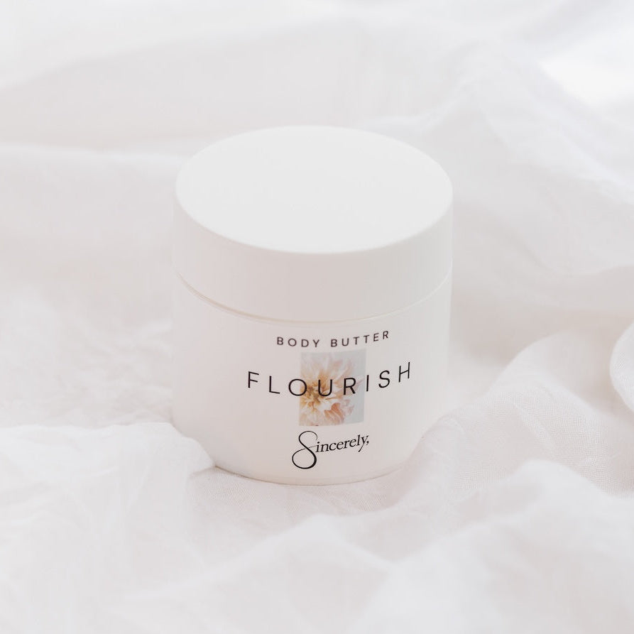 A white jar of Sincerely body butter labeled 'Flourish' sits on soft white draped fabric.