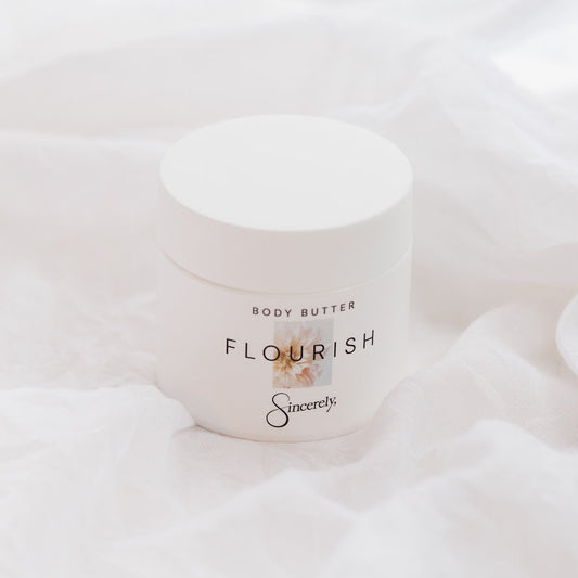 A white jar of Sincerely body butter labeled 'Flourish' sits on soft white draped fabric.