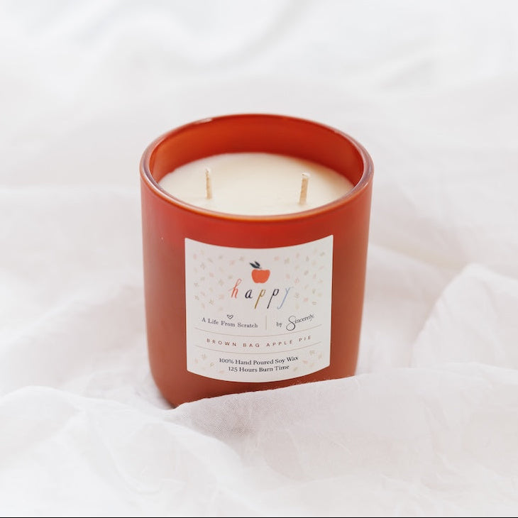 An orange-brown glass jar candle with cream-colored wax and a white label reading 'happy' and brown bag apple pie, set against a draped white fabric background.