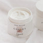 An open white jar of Sincerely Chalet body butter, showing the white cream inside, set against a soft white fabric background with another jar partially visible in the background.