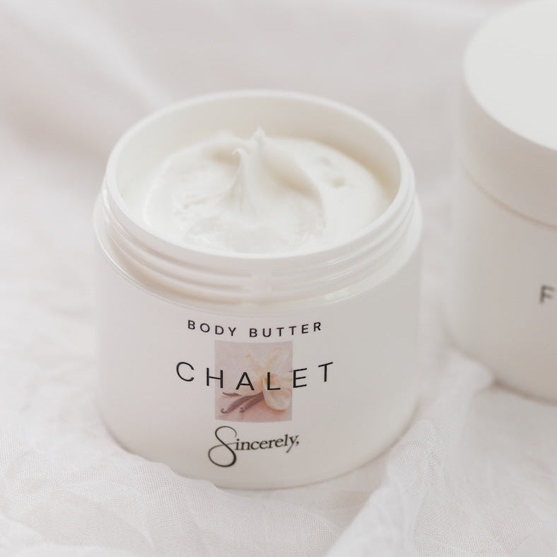 An open white jar of Sincerely Chalet body butter, showing the white cream inside, set against a soft white fabric background with another jar partially visible in the background.