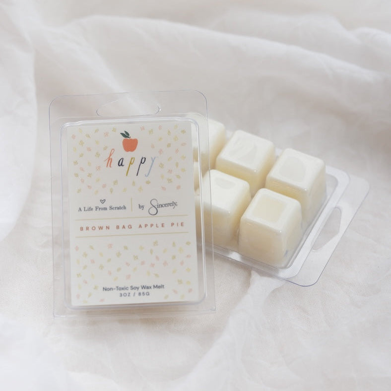 A clear plastic clamshell package of wax melts sits on draped white fabric, with several off-white wax cubes visible in the tray. The front label reads 'happy' with a small orange fruit icon and 'Brown Bag Apple Pie' scent text.