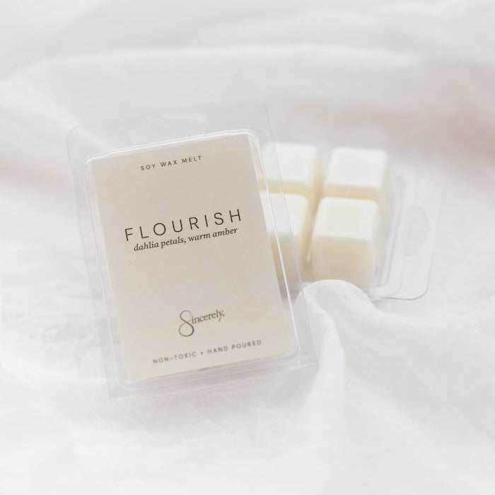A clear plastic clamshell package of wax melts labeled 'FLOURISH' with several off-white wax cubes visible, placed on soft white fabric.