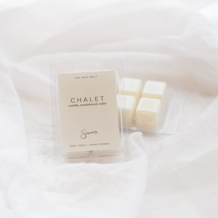 A clear plastic clamshell package of Chalet scented wax melts sits on draped white fabric next to three individual cream-colored wax cubes in a clear tray.
