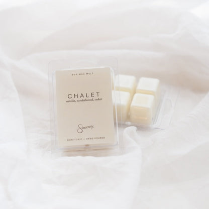 A clear plastic clamshell package of Chalet scented wax melts sits on draped white fabric next to three individual cream-colored wax cubes in a clear tray.
