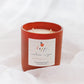 A red-orange glass jar candle with a white label reading 'happy by Brown Bag Apple Pie photographed on soft white fabric.