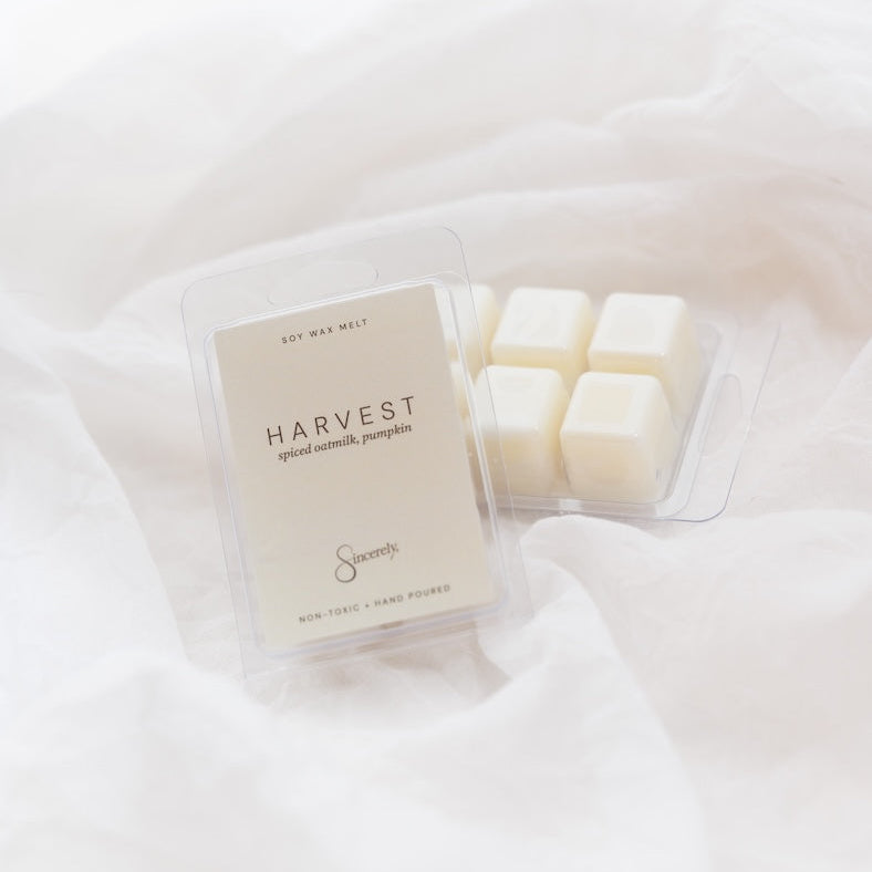A clear plastic clamshell package of Harvest scented wax melts sits on draped white fabric, with several cream-colored wax cubes visible inside the tray.