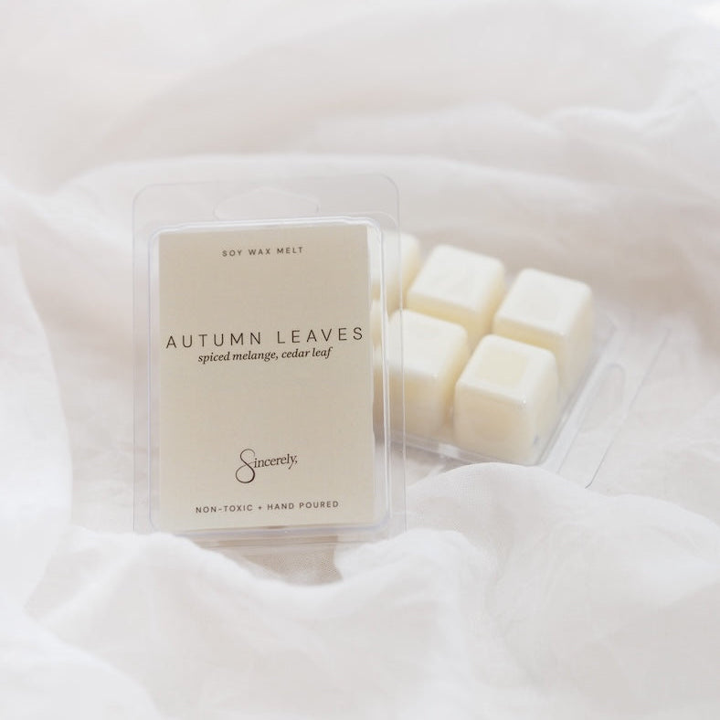 A clear clamshell package labeled 'Autumn Leaves' soy wax melt sits on soft white fabric, with several off-white wax cubes visible inside a clear tray behind it.