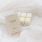 A clear plastic clamshell package of Hygge brown sugar wax melts sits on soft white fabric, with several cream-colored wax cubes visible inside the tray.