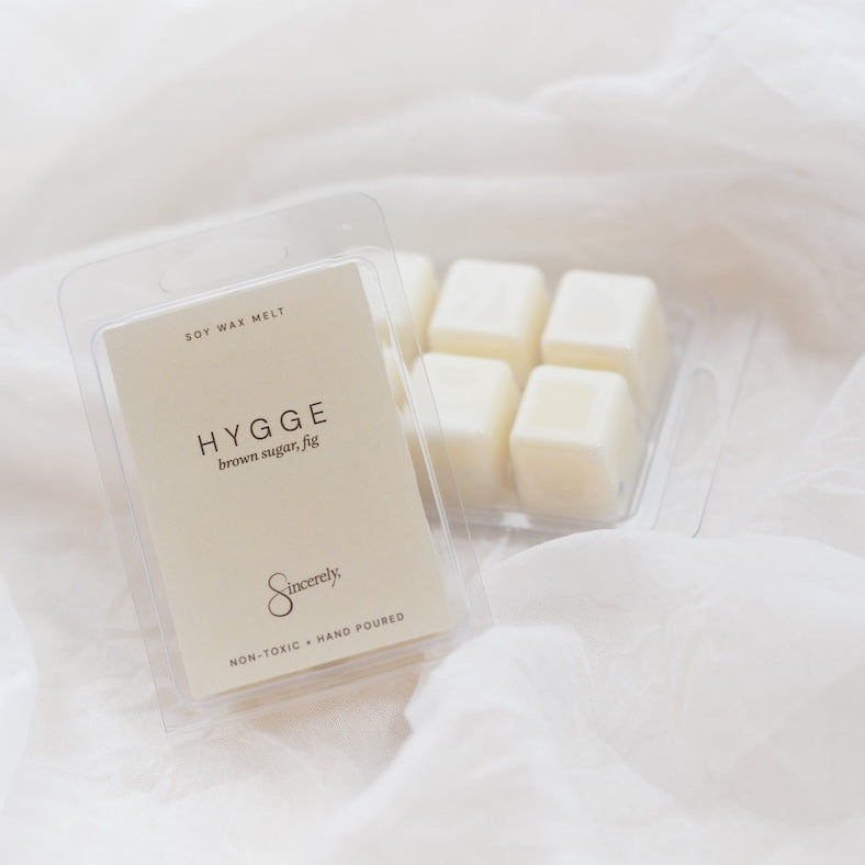 A clear plastic clamshell package of Hygge brown sugar wax melts sits on soft white fabric, with several cream-colored wax cubes visible inside the tray.