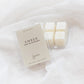 A clear plastic clamshell package of Ember soy wax melts with a cream-colored label, shown next to four white wax cubes in a matching clear container on a soft white fabric background.