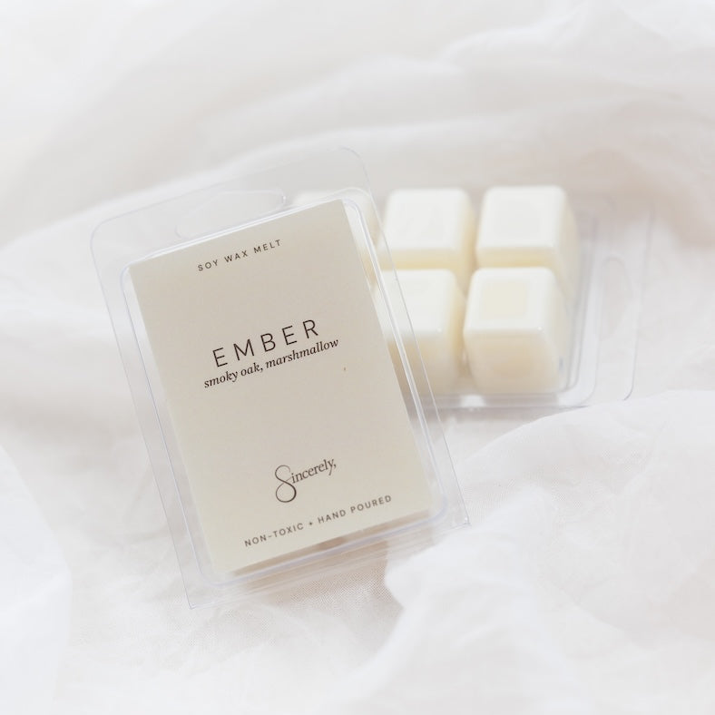 A clear plastic clamshell package of Ember soy wax melts with a cream-colored label, shown next to four white wax cubes in a matching clear container on a soft white fabric background.