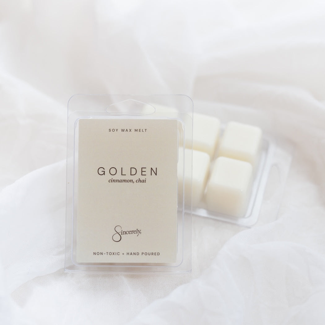 A clear plastic clamshell package of wax melts with a cream-colored label reading 'GOLDEN cinnamon, chai', placed on soft white fabric next to another wax melt package.
