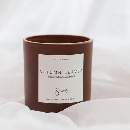 A brown soy candle jar with a white label reading 'AUTUMN LEAVES' and the brand name 'Sincerely' on a soft white fabric background.