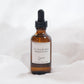 An amber glass dropper bottle of Flourish with a black dropper cap and a white label, set against a soft white fabric background.
