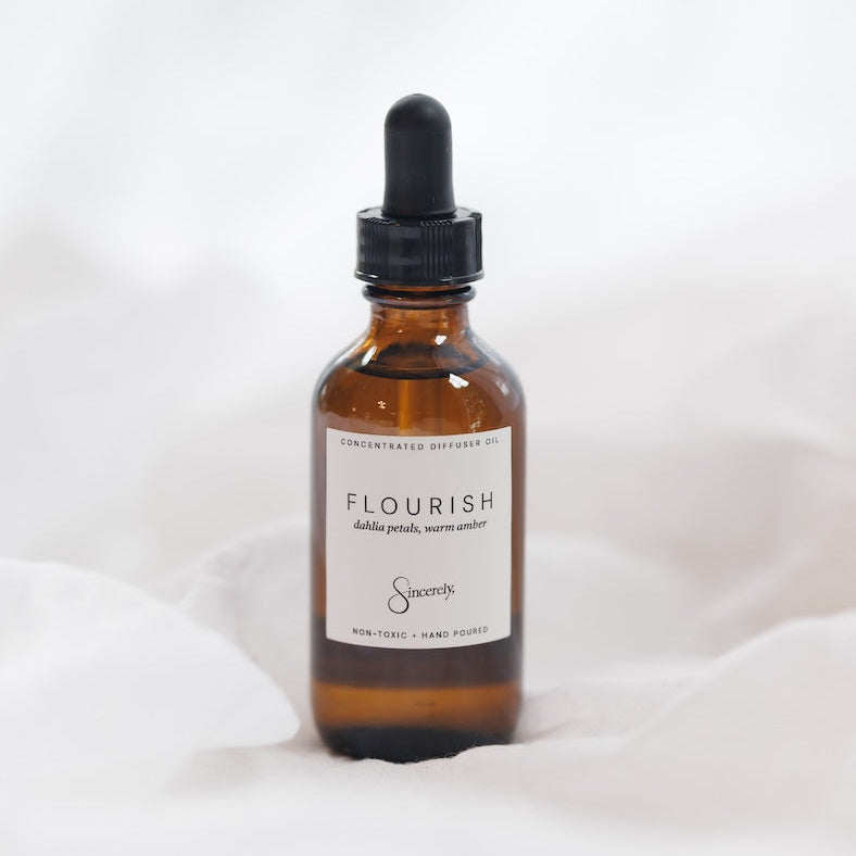 An amber glass dropper bottle of Flourish with a black dropper cap and a white label, set against a soft white fabric background.