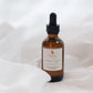 An amber glass dropper bottle with a black rubber bulb and a cream-colored label, set against a soft, draped white fabric background.
