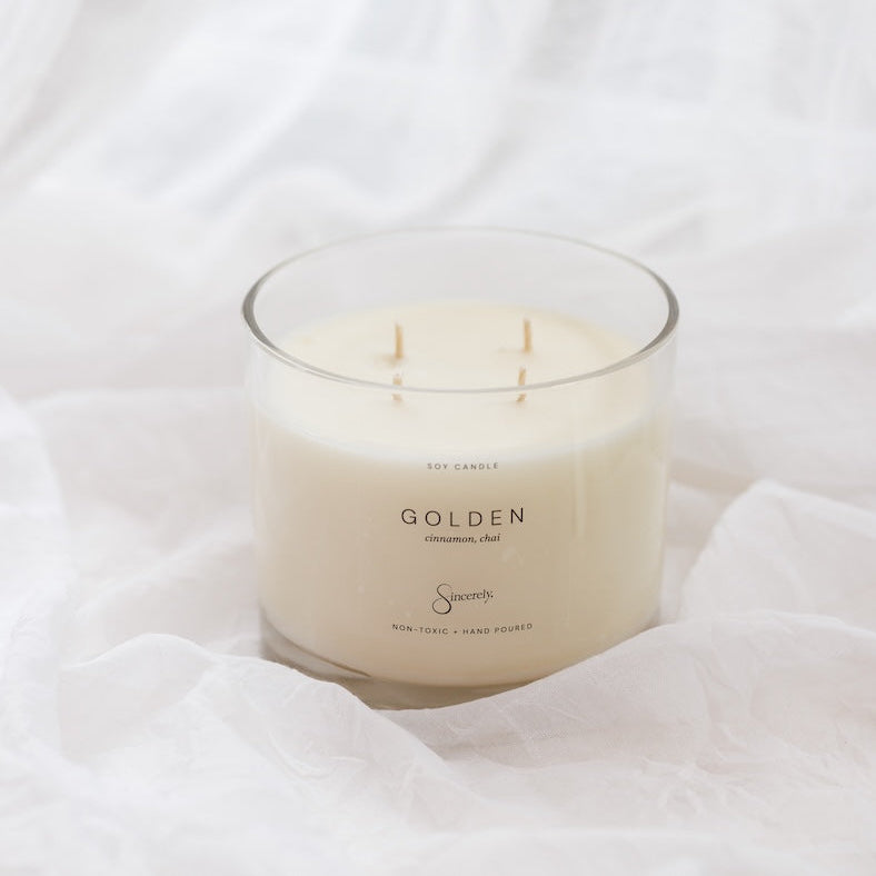 A 4-wick soy candle in a clear glass jar with cream-colored wax, labeled 'GOLDEN', set against a draped white fabric background.