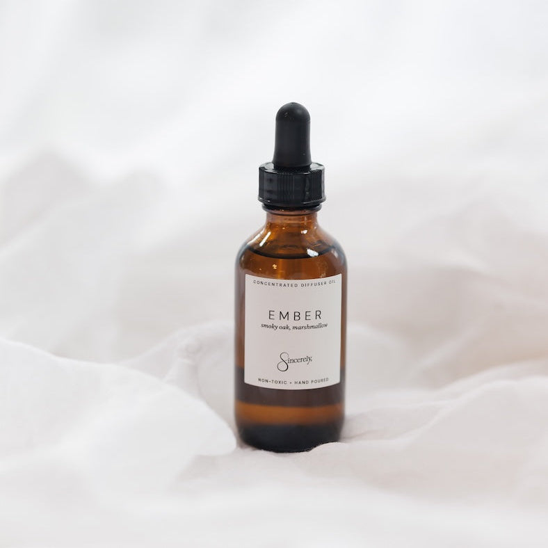 An amber glass dropper bottle with a black cap and a white label reading 'EMBER', set against a soft white draped fabric background.
