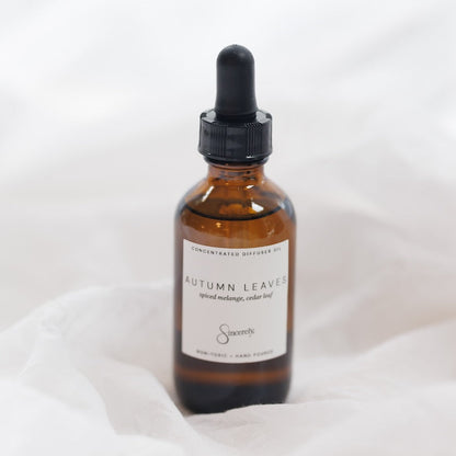 An amber glass dropper bottle with a black cap and a white label reading 'Autumn Leaves' and 'Sincerely', set against a soft white fabric background.