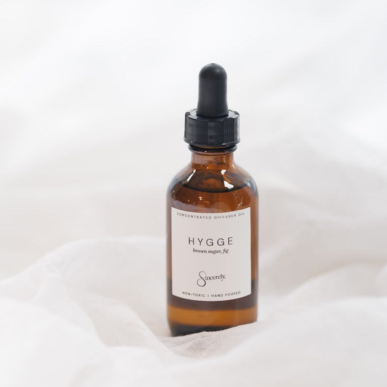 A small amber glass dropper bottle with a black cap and a cream label reading 'HYGGE', placed on soft white fabric against a white background.