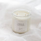 A soy candle in a clear glass jar with a white label reading 'EMBER', set on white fabric.