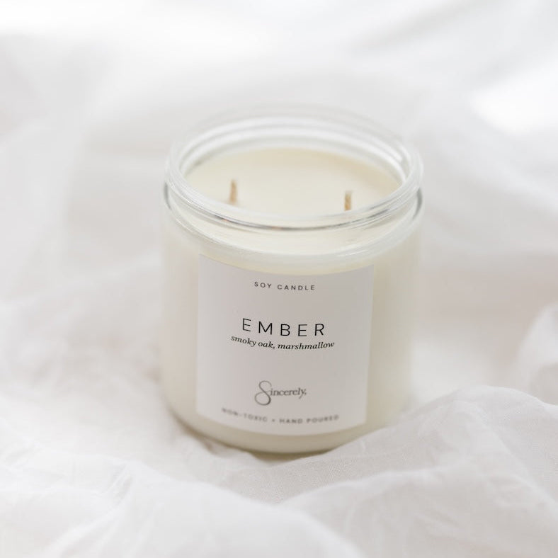 A soy candle in a clear glass jar with a white label reading 'EMBER', set on white fabric.