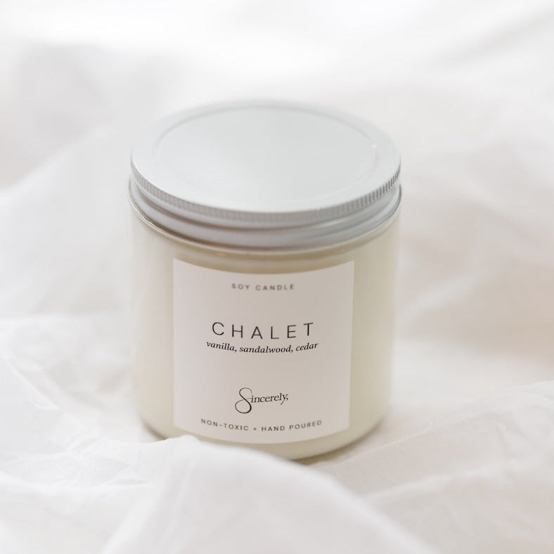 A soy candle in a clear glass jar with a white metal lid and a white label reading 'CHALET', set against a soft white fabric background.