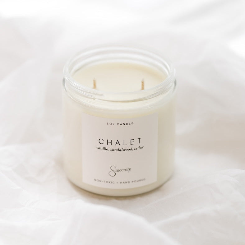 A two-wick soy candle in a clear glass jar with a cream-colored label reading 'CHALET', set against a softly draped white fabric background.