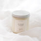 A soy candle in a clear glass jar with a white lid and a minimalist white label reading 'HYGGE' set against a soft white draped fabric background.