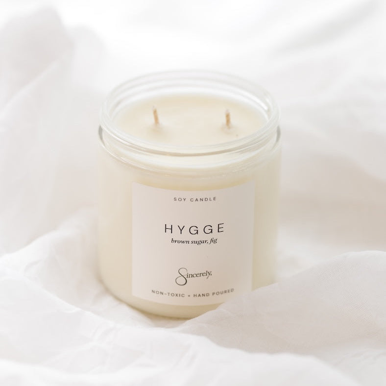 A two-wick soy candle in a clear glass jar with a cream-colored label reading 'HYGGE' and 'Sincerely', styled on draped white fabric.