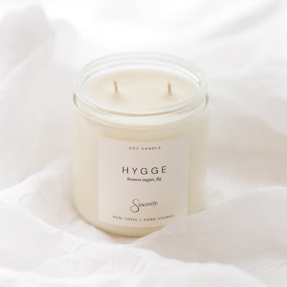 A two-wick soy candle in a clear glass jar with a cream-colored label reading 'HYGGE' and 'Sincerely', styled on draped white fabric.