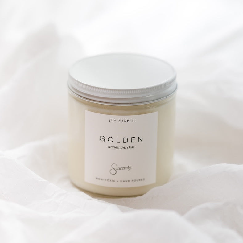 A soy candle in a clear glass jar with cream-colored wax and a white label reading 'GOLDEN cinnamon, chai, set against a soft, draped white fabric background.