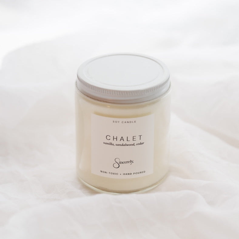 A soy candle in a clear glass jar with a white lid and a cream-colored label reading 'CHALET' set against a soft white fabric background.