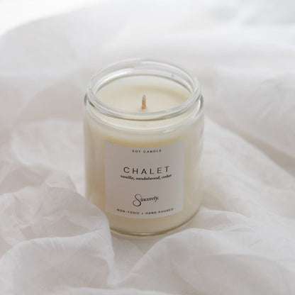 A soy candle in a clear glass jar with a cream-colored wax and a white label reading 'CHALET', set on a soft, draped white fabric background.