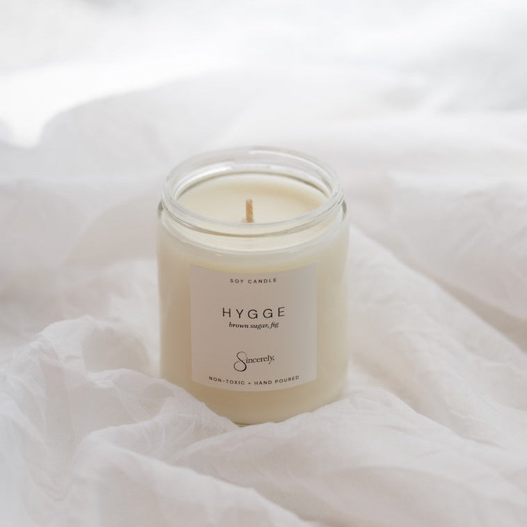 A glass jar candle with cream-colored wax sits on draped white fabric. The front label reads 'HYGGE' with smaller text below and a script brand mark near the bottom.