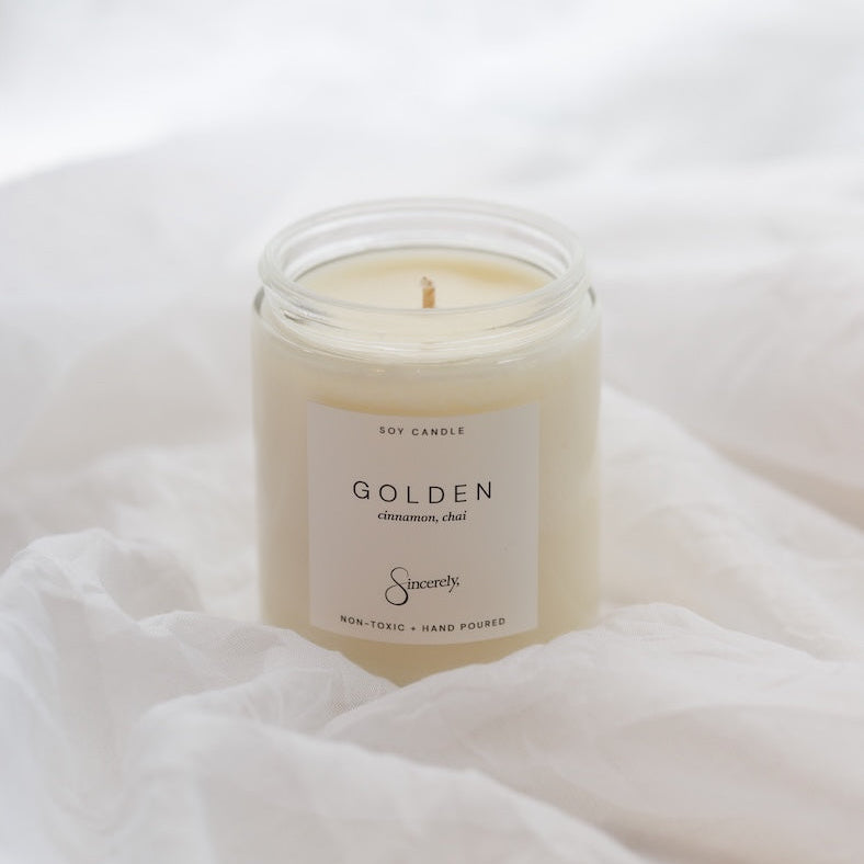 A soy candle in a clear glass jar with a white label reading 'GOLDEN' and 'Sincerely', placed on soft white fabric.