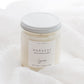 A soy candle in a clear glass jar with a white lid and a white label reading 'HARVEST' set against a soft white fabric background.