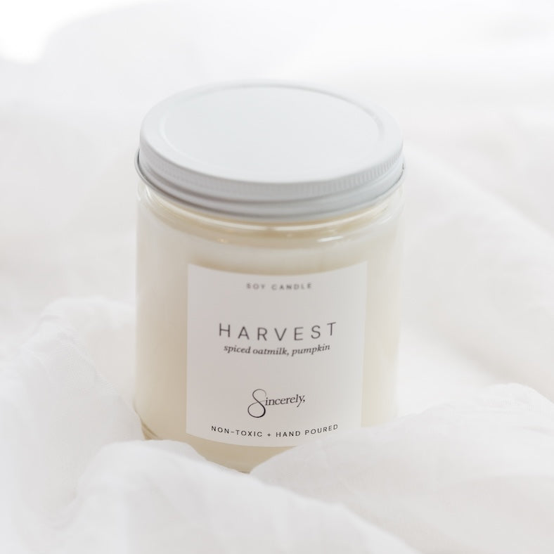 A soy candle in a clear glass jar with a white lid and a white label reading 'HARVEST' set against a soft white fabric background.