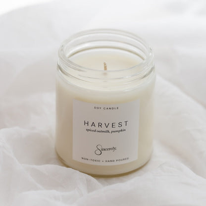 A soy candle in a clear glass jar with a white label reading 'HARVEST' and 'spiced oatmilk and pumpkin' placed on soft white fabric.