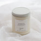 A jar candle labeled 'EMBER' by Sincerely, placed on soft white fabric. The candle has a white wax fill, a white label, and a white metal screw lid.