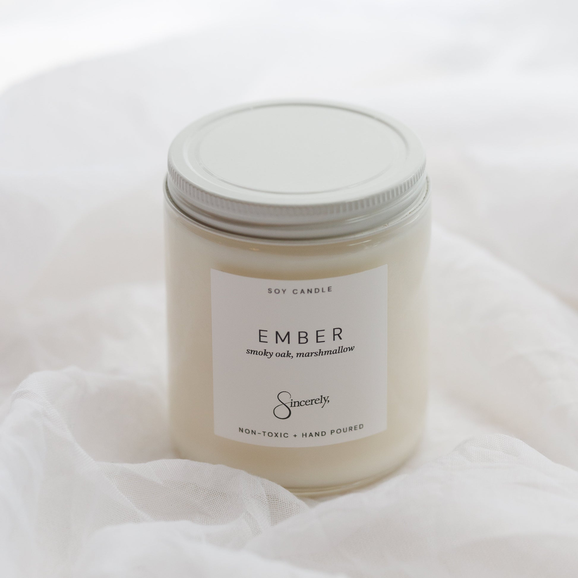 A jar candle labeled 'EMBER' by Sincerely, placed on soft white fabric. The candle has a white wax fill, a white label, and a white metal screw lid.