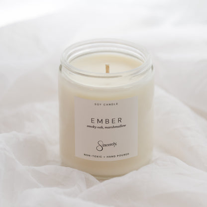 A jar candle labeled 'EMBER' set on draped white fabric. The candle is in a clear glass jar with creamy off-white wax and a white label.