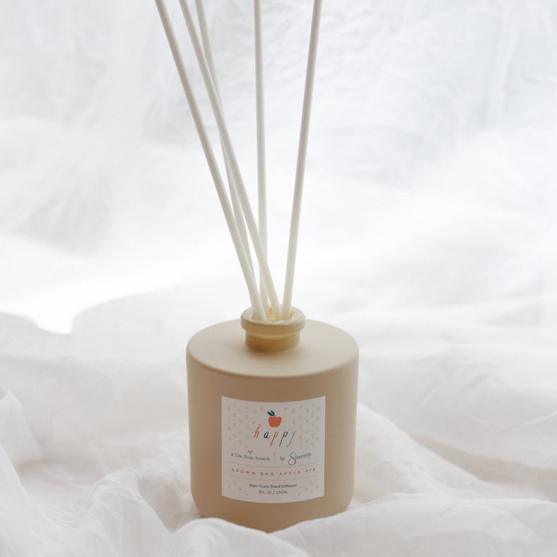 A reed diffuser bottle in a matte beige finish with natural reed sticks, resting on a soft white fabric background.