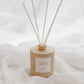 A reed diffuser bottle in a warm beige tone with four natural reed sticks, set against a soft white draped fabric background. The label reads 'EMBER'