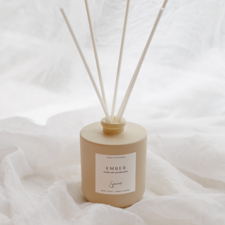 A reed diffuser bottle in a warm beige tone with four natural reed sticks, set against a soft white draped fabric background. The label reads 'EMBER'