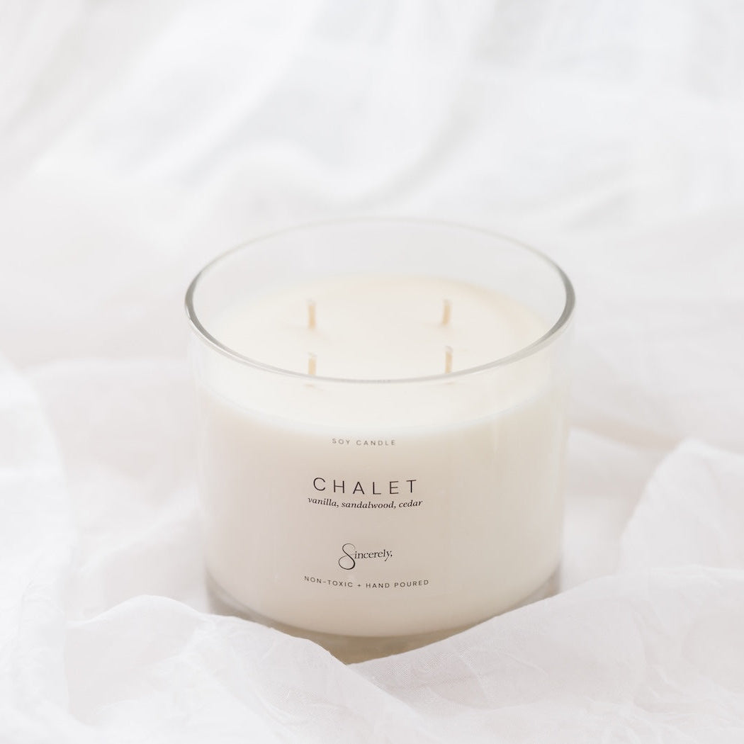 A 4-wick soy candle in a clear glass jar with white wax, labeled 'CHALET', set against a background of soft, draped white fabric.