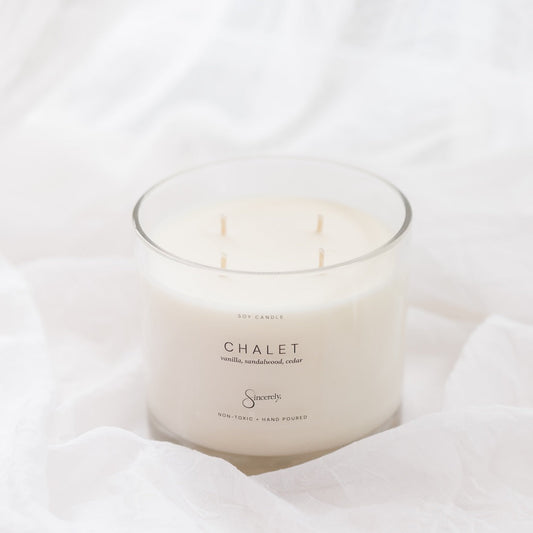 A 4-wick soy candle in a clear glass jar with white wax, labeled 'CHALET', set against a background of soft, draped white fabric.